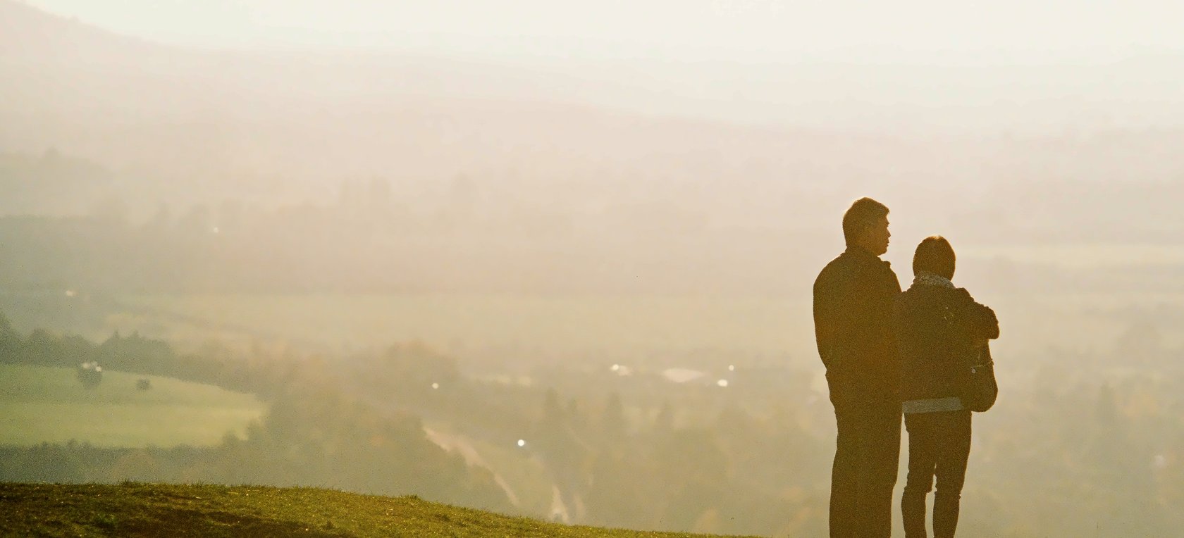 Two people standing on a hillside, looking out over a hazy valley in the early morning
